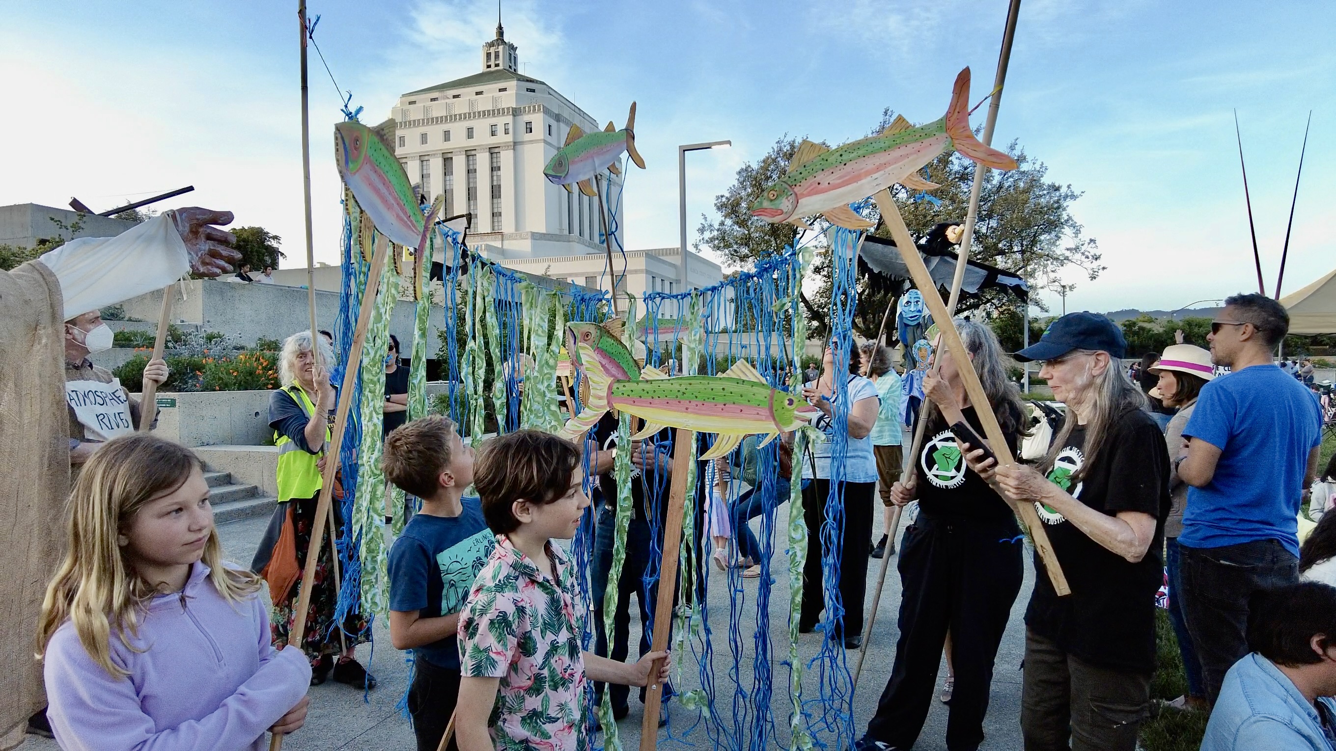 Youth and adults stand in a plaza holding blue and green banners and painted salmon